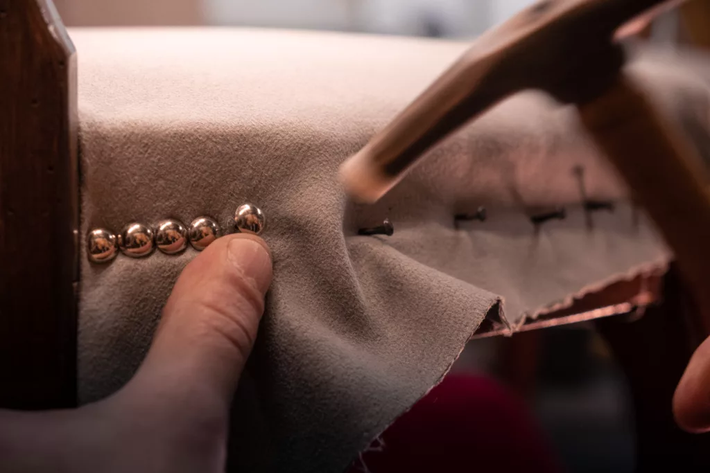 travail à la main avec un ramponneau permettant d'insérer des clous décoratifs dorés sur une assise de siège de couleur beige